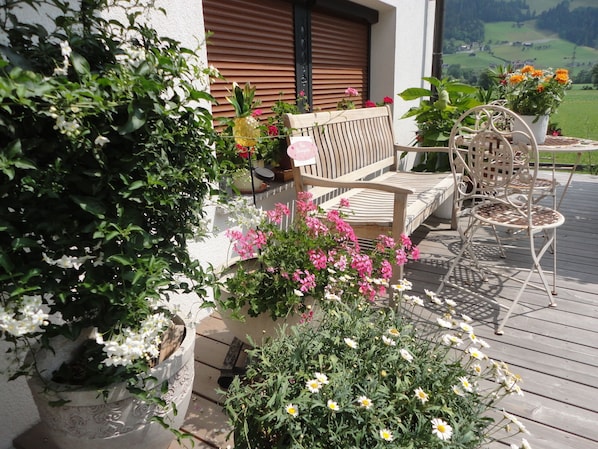 Um deck de madeira com um banco e cadeiras, cercado por plantas e flores em vasos.
