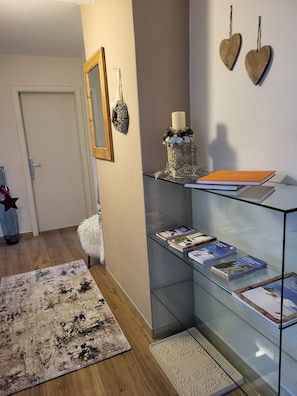 Um corredor com uma vitrine de vidro contendo livros, uma poltrona branca e fofa, uma estrela de madeira decorativa e um espelho na parede.