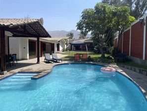 En pool med solstolar, en möblerad sittgrupp under tak och utsikt över berg i bakgrunden.