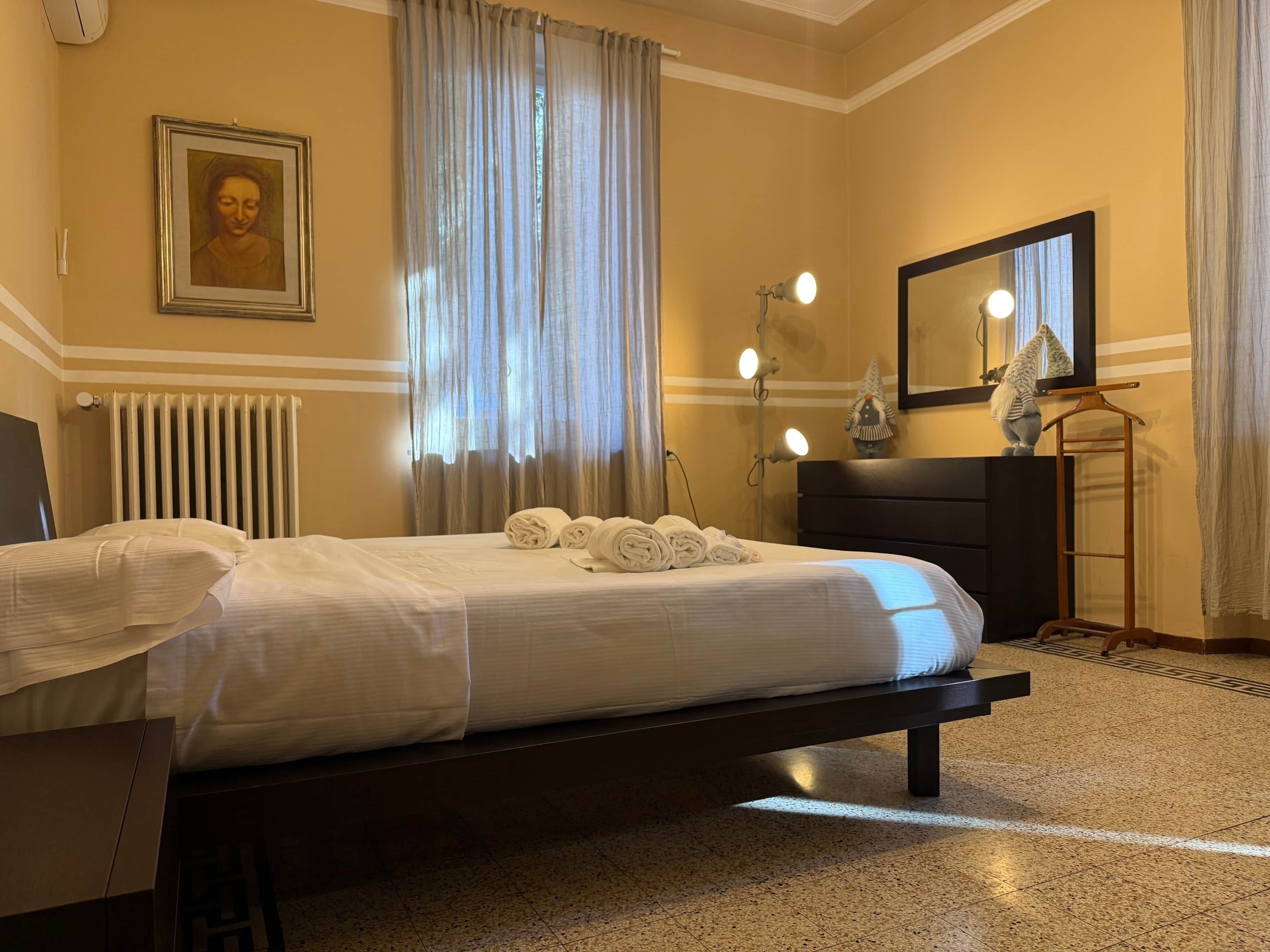 Una habitació d'hotel amb llit, radiador, finestra amb cortines, mirall i una petita estàtua a la petita taula de nit.