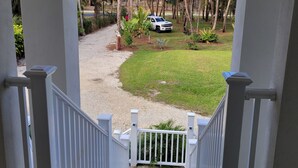 A wooden staircase leading to a porch with white railings, overlooking a well-maintained lawn with palm trees and a white vehicle parked nearby. - Pine Ridge Model Home (Naples)