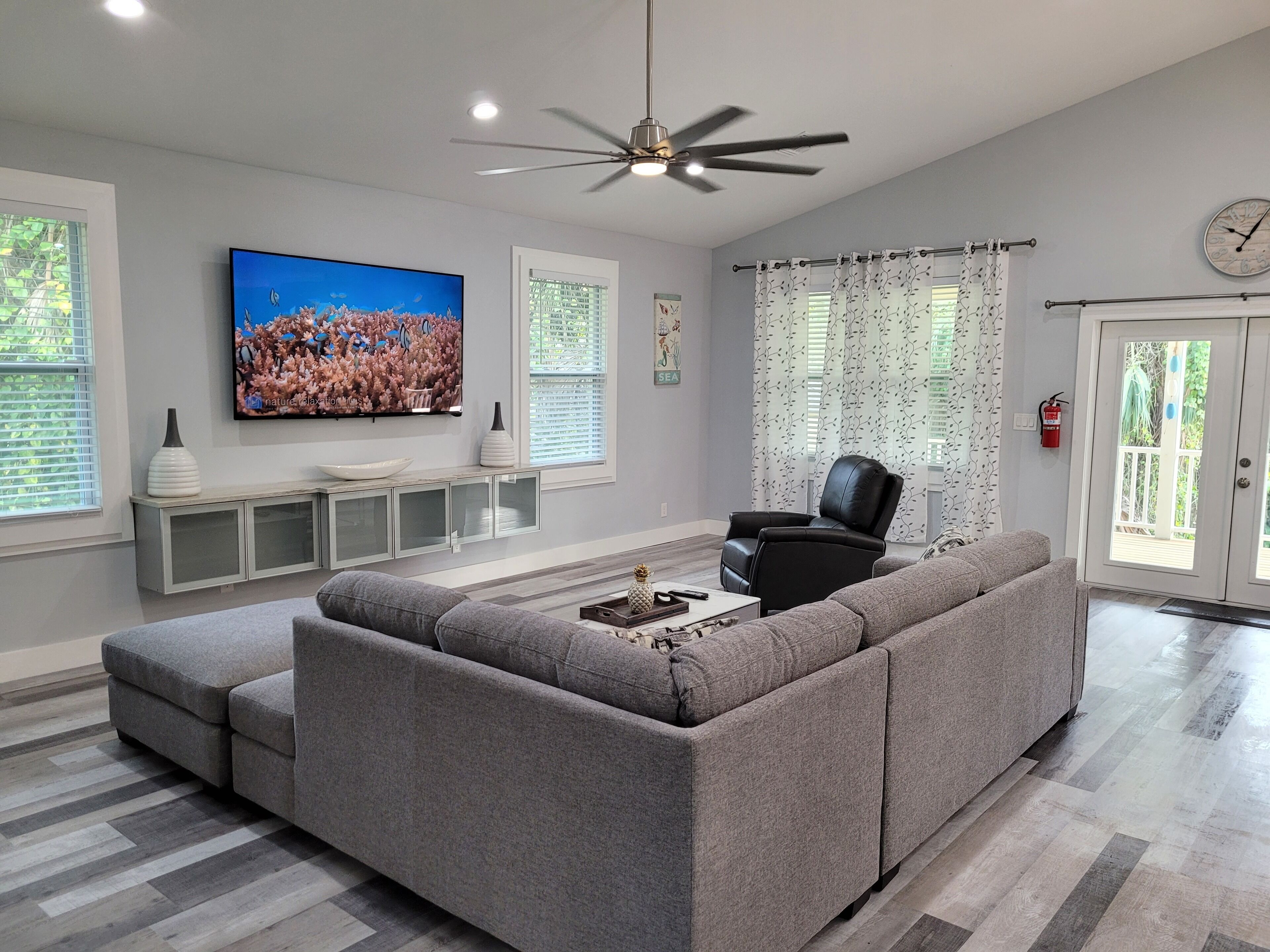 A modern living room with a grey sectional sofa, a flat-screen TV, a ceiling fan, and a large window with curtains.