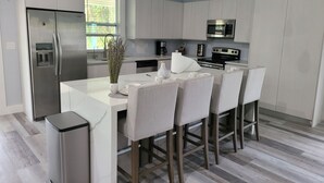 A modern kitchen with white cabinetry, a stainless steel refrigerator, a microwave, and a dining area with bar stools. - Pine Ridge Model Home (Naples)