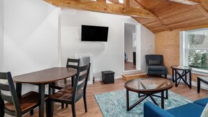 Un interior moderno con comedor, sala de estar con sofá y mesa de centro, televisor montado en la pared y un amplio ventanal que proporciona luz natural.