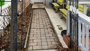 Un estrecho sendero pavimentado que conduce a una entrada con puerta de reja y valla de piquetes blancos, con sillas amarillas.