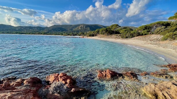 Ein Küstenstrand mit klarem türkisblauem Wasser, felsigem Ufer und grünen Hügeln im Hintergrund.