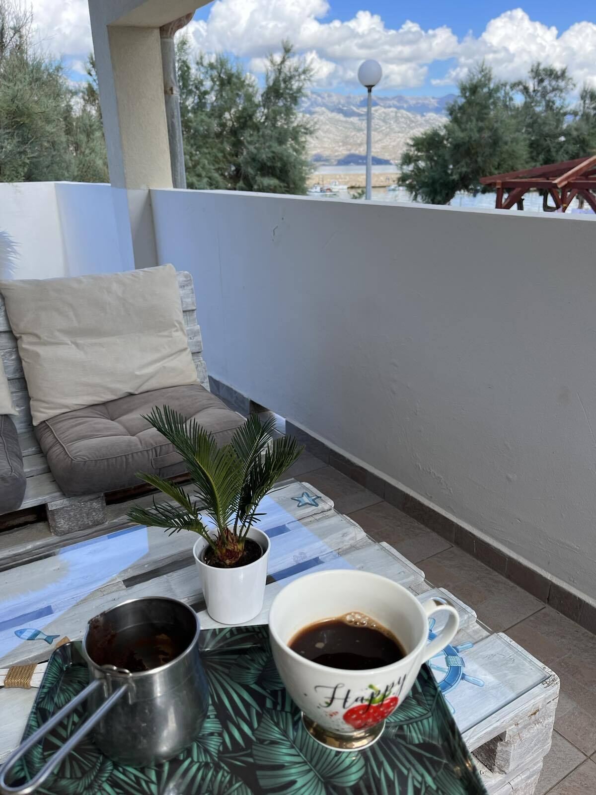 A balcony with a view of mountains, a potted plant, a metal cup, and a white mug with a design.
