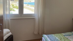 Quarto com piso de madeira, cama com colcha verde e branca, janela com cortinas brancas e vista para uma praia e edifícios.