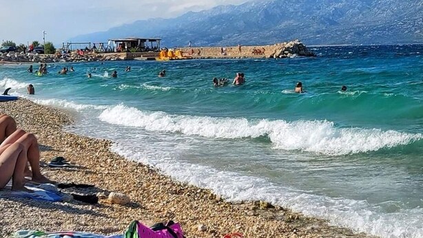 Uma praia com pessoas nadando e tomando sol, um litoral rochoso e montanhas ao fundo.