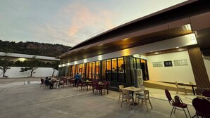 An outdoor café area with tables and chairs, a building with large windows, and a view of a hilly landscape. - P.T. Grand Hotel (Kanchanaburi)