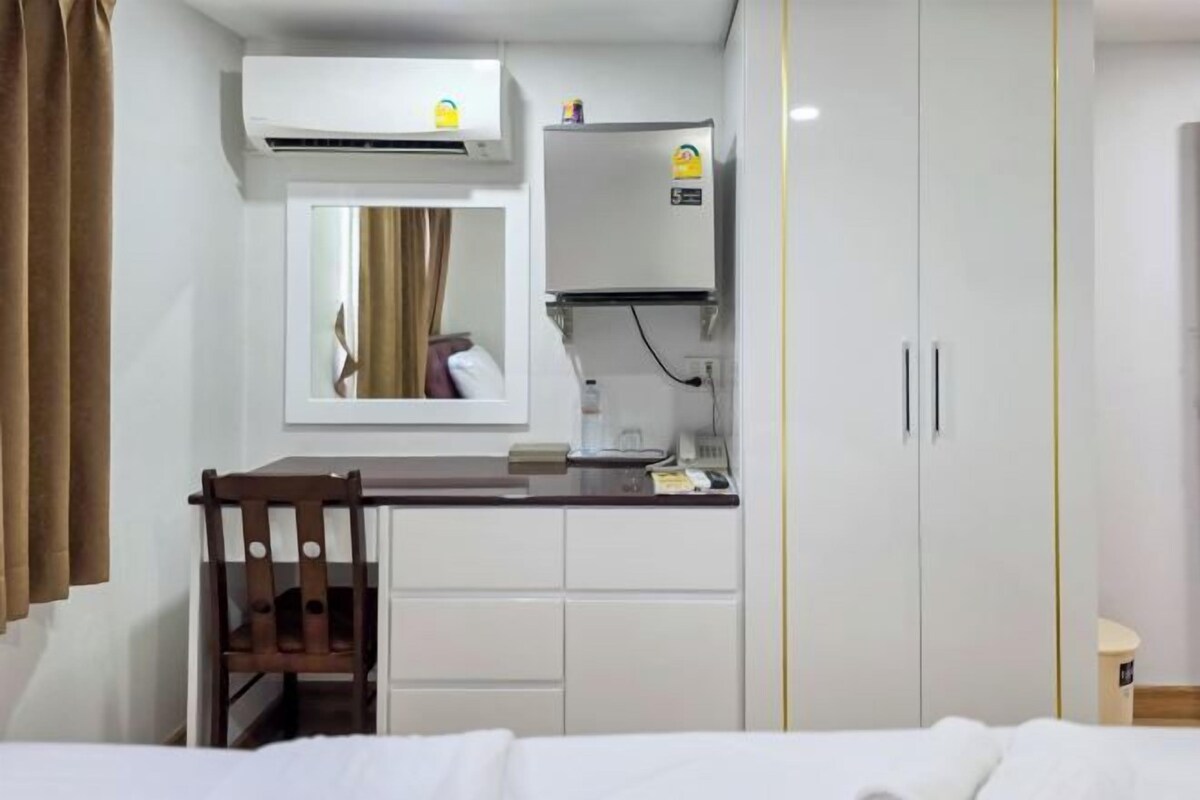 A compact hotel room with a bed, a small desk with a chair, a wardrobe, an air conditioning unit, and a mirror.