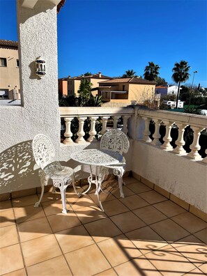 Un balcone con tavolo e sedia in ferro battuto bianco, pavimento piastrellato e vista su edifici e palme circostanti.