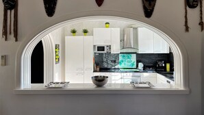 Une cuisine avec des meubles blancs, un four à micro-ondes et une cuisinière. Des masques décoratifs sont accrochés au mur, au-dessus de la fenêtre.