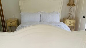 A white ornate bed with a curved headboard, two bedside tables with drawers, a lamp, and a mirror on the wall.