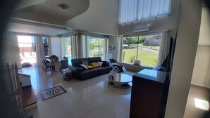 A spacious living room with a black leather sofa, a coffee table, and a TV. There are large windows providing a view of the outdoors. - Beautiful house in Campeche for up to 16 people, heated pool and spa. (Florianópolis)