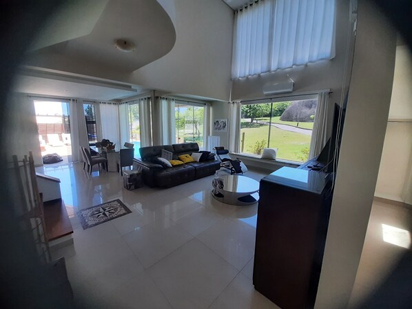 A spacious living room with a black leather sofa, a coffee table, and a TV. There are large windows providing a view of the outdoors.