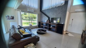 A modern living room with a brown leather sofa, a TV on a wooden stand, and a view of a green landscape through large windows. - Beautiful house in Campeche for up to 16 people, heated pool and spa. (Florianópolis)