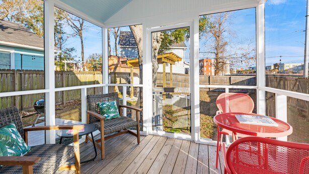 Una terraza techada al aire libre con sillas de mimbre y metal, una mesa y vistas a un patio trasero con cerca de madera y una casa.