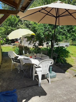 An outdoor patio with a table set for dining, surrounded by white plastic chairs, under a large umbrella.