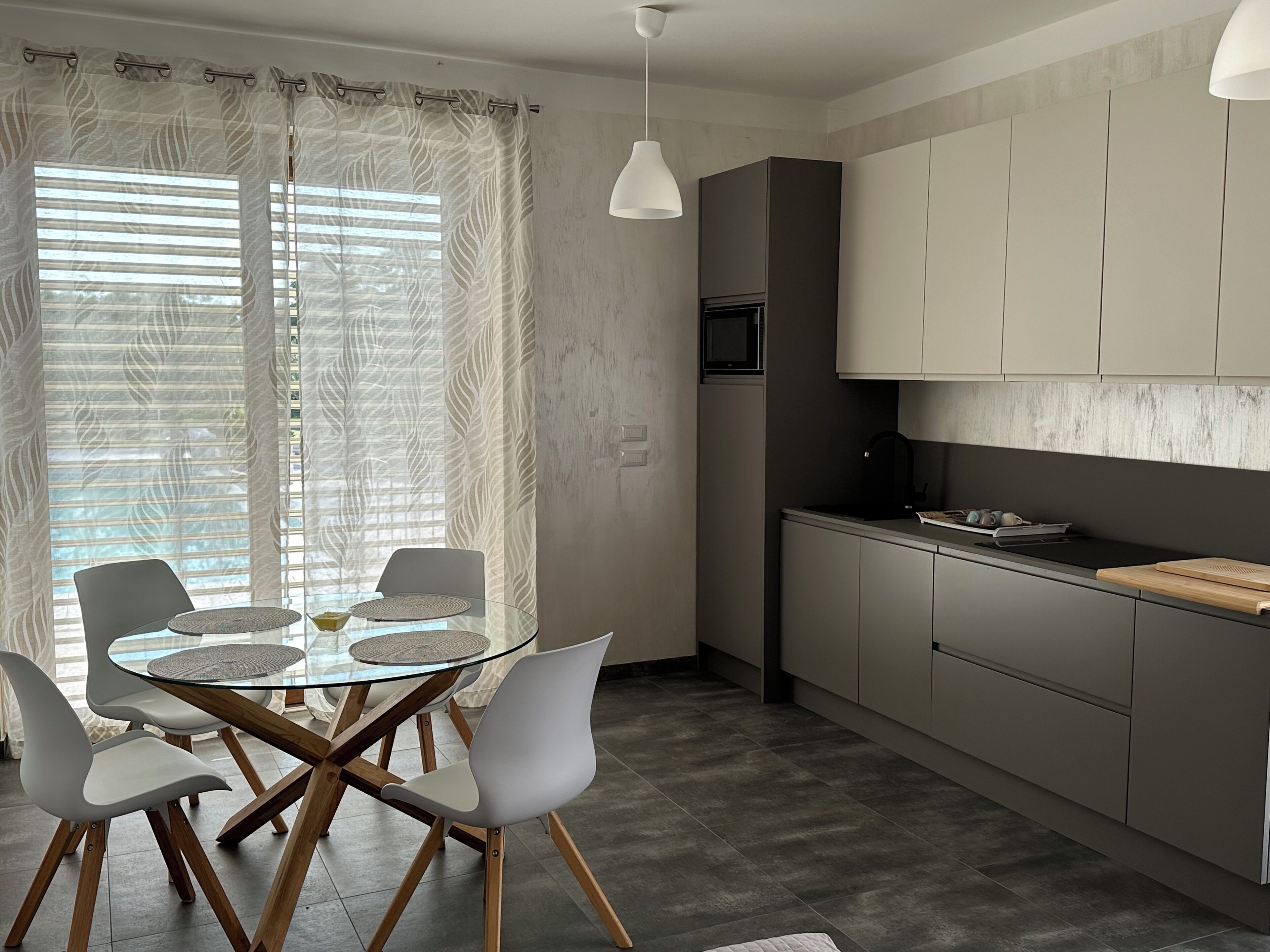 A modern kitchen with a dining area, featuring a glass-top table, white chairs, and grey cabinets.