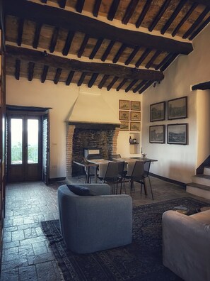 Un interno rustico con travi a vista in legno, un camino in pietra, un'area pranzo con tavolo e sedie, e un'ampia finestra con vista.