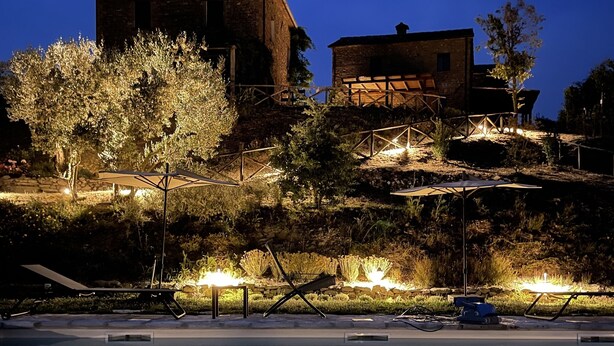 Une vue nocturne d’une piscine aux carreaux de pierre, d’une terrasse en bois et d’un bâtiment à l’aspect rustique.