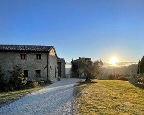Un edificio in pietra con un vialetto sterrato che vi conduce, circondato da erba e alberi.