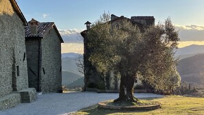 Un bâtiment en pierre avec un toit recouvert de tuiles, un arbre au premier plan et un paysage montagneux en arrière-plan.