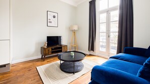 A modern living room with a blue sofa, a round black coffee table with a plant, a wooden side table with a TV, and a floor lamp. - The Chelsea Collection (London)