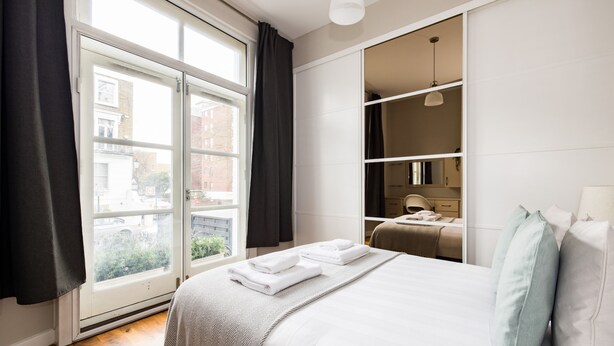 A modern hotel room with a large window, a bed with neatly folded white towels, a wardrobe, and a desk with a lamp. - The Chelsea Collection (London)