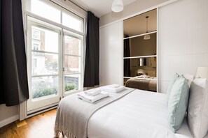 A modern hotel room with a large window, a bed with neatly folded white towels, a wardrobe, and a desk with a lamp. - The Chelsea Collection (London)