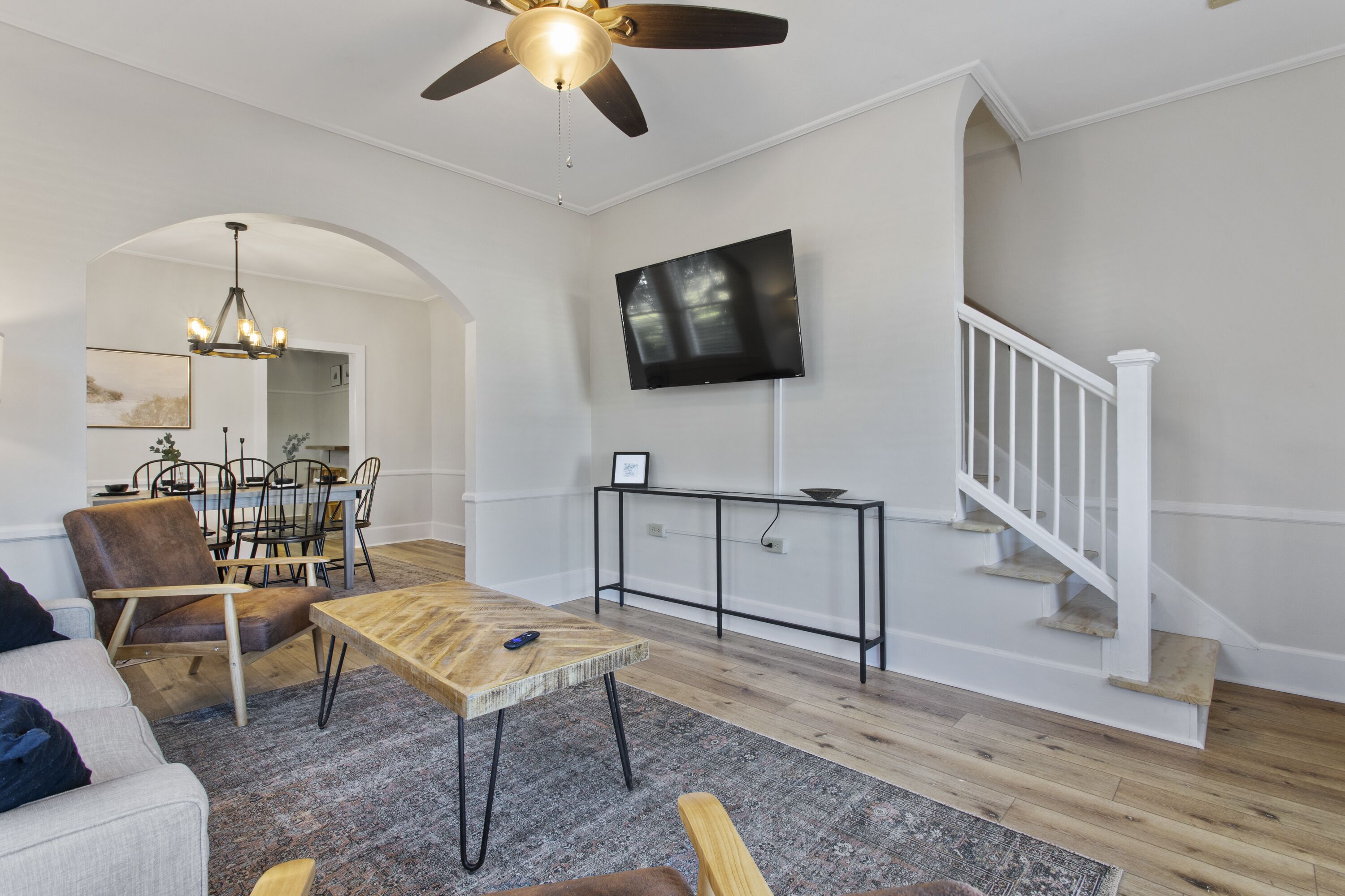 Un salón amplio con piso de madera, una zona de comedor con mesa y sillas, un televisor montado en la pared y una escalera con barandales blancos.