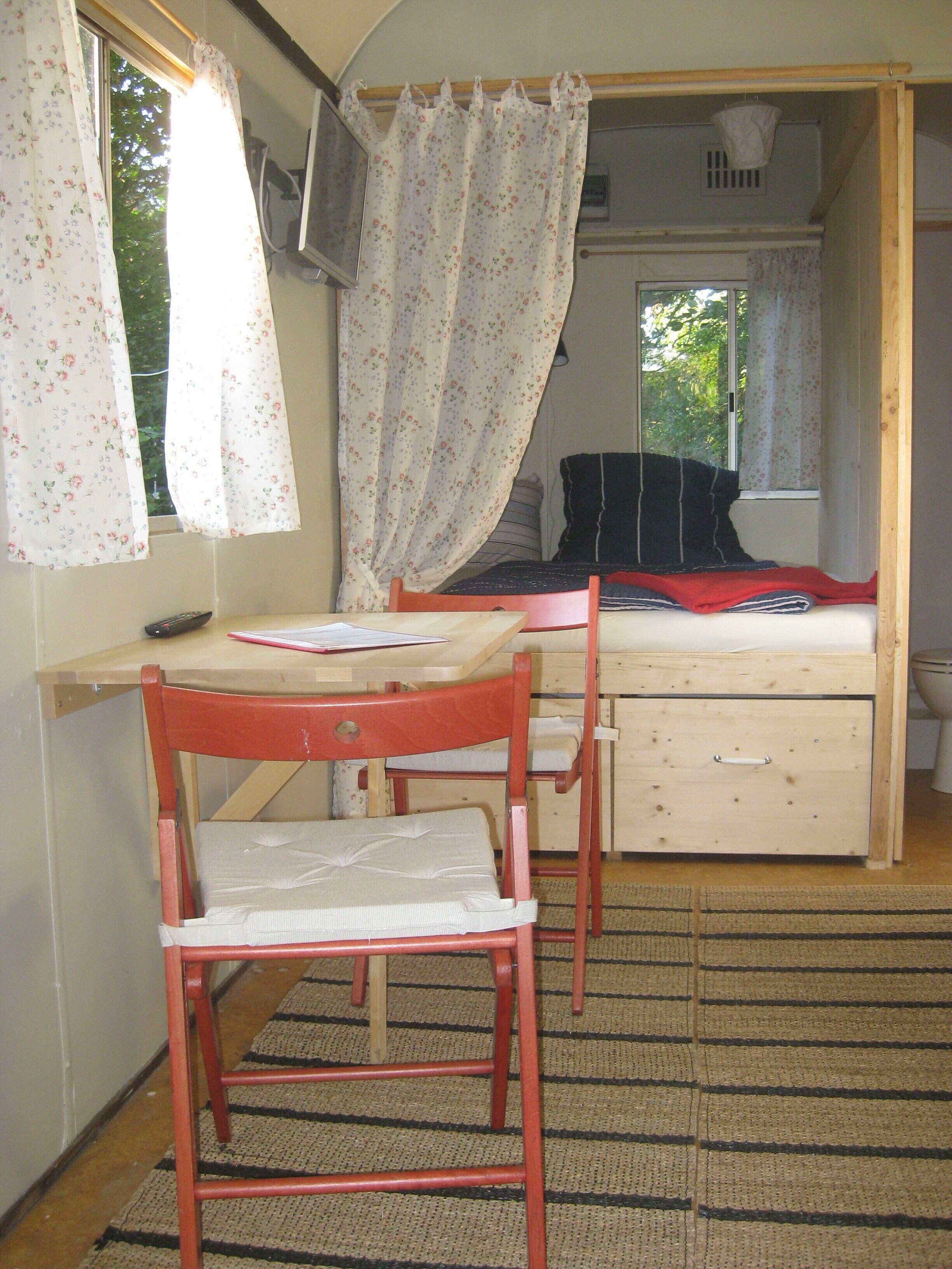 A small bedroom with a single bed, a wooden chair, a desk, and a window with floral curtains.