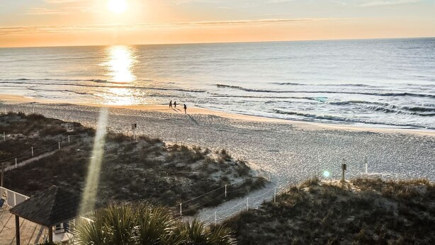 Una playa al atardecer, con gente caminando y una cerca en primer plano.