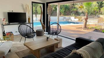 Un salon moderne avec un téléviseur, deux fauteuils noirs, une table en bois et un canapé. Une grande fenêtre offre une vue sur une piscine et sur la verdure environnante.