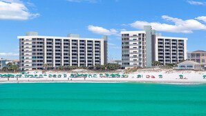 Ein Strandhotel mit mehreren Etagen, Balkonen und einem Sandstrand mit Sonnenschirmen.