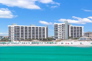 A beachfront hotel with multiple floors, balconies, and a sandy beach area with umbrellas.