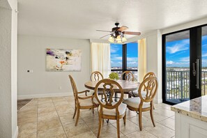 A dining area with a round table, six wooden chairs, a ceiling fan, a large window with a view, and a painting on the wall.