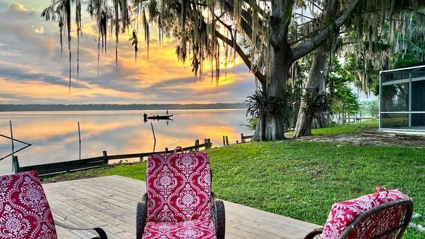 A wooden deck with wicker chairs, a view of a lake, and a large tree with hanging moss. - Peaceful & stylish pool home with gorgeous lake views (Lake Panasoffkee)