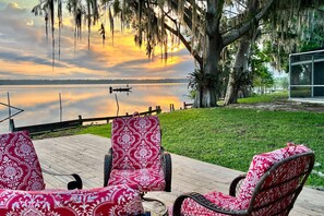 Une terrasse en bois avec des chaises en osier, une vue sur un lac et un grand arbre recouvert de mousse pendant.