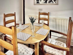 A round wooden dining table with four chairs, each with a white cushion. A vase with a plant is on the table, and there are two wine glasses.