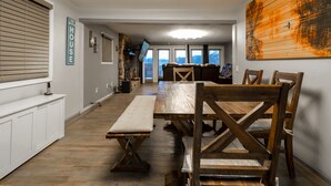 Un comedor amplio con una larga mesa de madera y bancos, una pared blanca con el letrero 'The Beach House', y una gran obra de arte de madera en la pared.