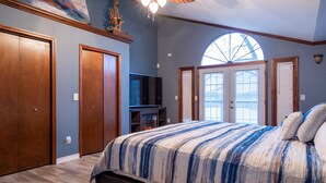 Un dormitorio con una cama grande, un televisor y una chimenea. Se ven dos puertas y una ventana con persianas.