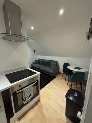 A compact living space with a kitchenette, a sofa, a small round table, and a trash bin.