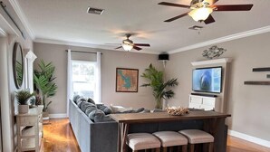 Ein modernes Wohnzimmer mit einem grauen Ecksofa, einem Holzesstisch mit Barhocker, einem Deckenventilator und einem Flachbildfernseher.