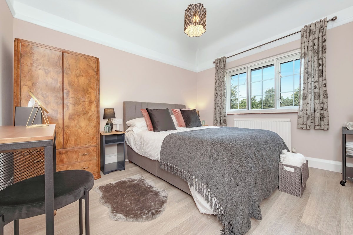 Quarto com cama, mesas de cabeceira, escrivaninha, cadeira, guarda-roupa, janela com cortinas e luminária pendente.