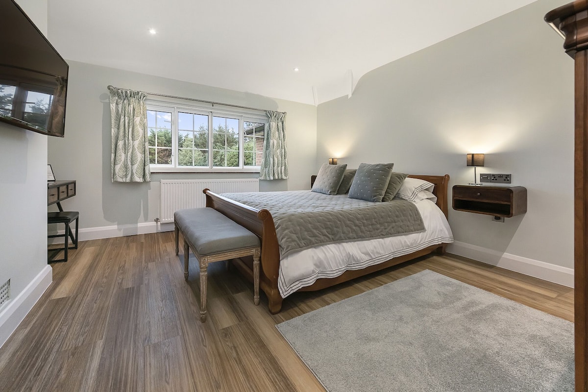 Quarto com cama, banqueta, mesa de cabeceira, luminária e vista para o exterior através de uma janela.