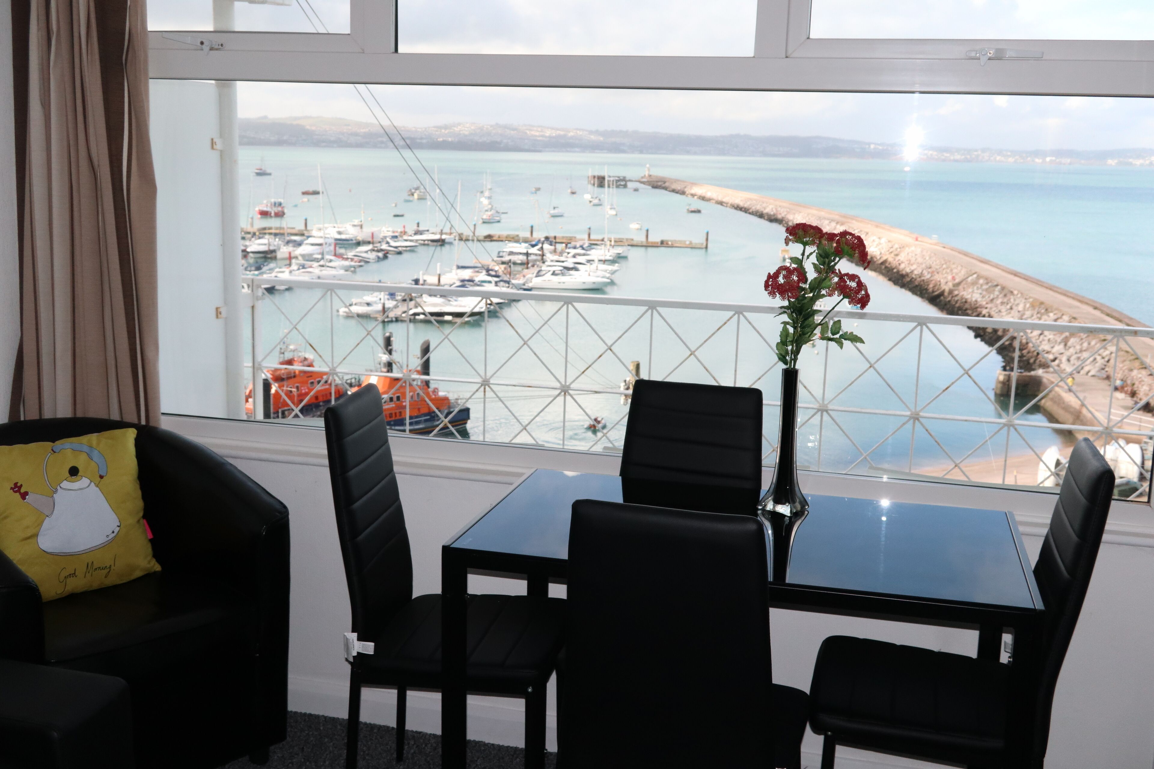 Een balkon met een tafel en stoelen, uitkijkend op een jachthaven met boten en een strand.