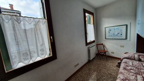 A small, sparsely furnished room with a large window, a radiator, and a framed picture on the wall.