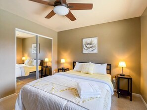 Une chambre d’hôtel comprenant un lit, deux tables de chevet avec des lampes, un ventilateur de plafond, un grand miroir et une œuvre d’art accrochée au mur.